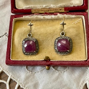 Peut inclure: Boucles d'oreilles en argent avec des pierres précieuses carrées, facettées, rouge foncé. Les pierres sont bordées de petites pierres claires. Les boucles sont présentées dans un écrin rouge avec un intérieur jaune.