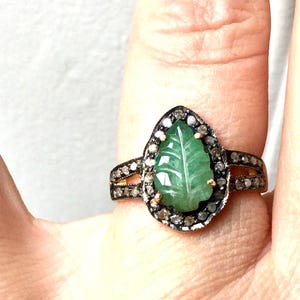Peut inclure: Bague en forme de feuille avec une pierre verte sculptée au centre, entourée de petites pierres scintillantes. La bague a un double anneau avec d'autres petites pierres, serties dans un métal doré.