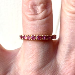 Peut inclure: Une bague en or ornée d'une rangée de pierres précieuses roses. La bague est présentée sur un doigt, les pierres précieuses captant la lumière. La bague est un design classique, adapté au quotidien ou aux occasions spéciales.