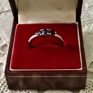 May include: A silver ring with three blue sapphires set in a pave setting. The ring is in a red velvet ring box.