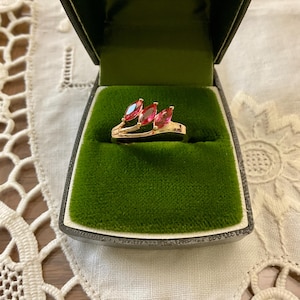 Peut inclure: Une bague en or avec trois pierres roses en forme de marquise serties dans une bande incurvée. La bague est dans un écrin à bague en velours vert avec le texte "3, Rue de la Fenderie CHARLEROI" sur le couvercle.