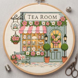 Op de afbeelding: Een kruissteekborduurwerk in een houten ring met een theesalon-winkelpui, een roze-wit gestreepte luifel, bloemdecoraties en de tekst TEA ROOM. De afbeelding bevat een etalage met theepotten, een deur en planten in potten.