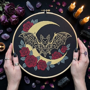 May include: A circular wooden embroidery hoop containing a cross-stitch design on black fabric. The pattern features a central gold crescent moon with a detailed gold bat silhouette in front, surrounded by deep red roses and grey-green leaves.