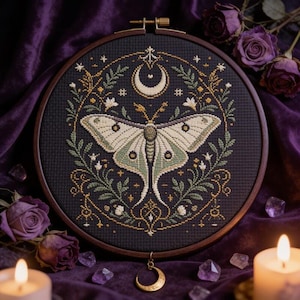 May include: A circular wooden embroidery hoop containing a cross-stitch design on black fabric. The central motif is a pale green and white moth surrounded by a circular wreath of green leaves, small white flowers, and a crescent moon at the top.