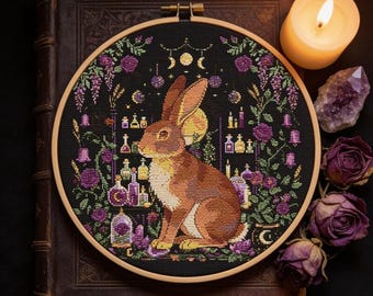 May include: A circular wooden embroidery hoop containing a cross-stitch design on black fabric. The central image is a brown rabbit surrounded by purple flowers, glowing potion bottles, and a moon phase motif. The design features vibrant colors.