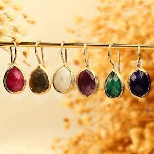 Könnte beinhalten: Sechs Paar vergoldete Ohrringe mit verschiedenen farbigen Edelsteintropfen. Die Ohrringe haben ein tropfenförmiges Design mit einer goldenen Fassung. Die Edelsteine sind rot, grün, lila, blau, weiß und schwarz.