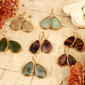 May include: A set of six pairs of teardrop-shaped gemstone earrings with gold-toned settings. The earrings feature a variety of gemstones, including amethyst, green aventurine, tiger's eye, rose quartz, aquamarine, and fluorite.