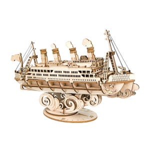 May include: A wooden model of a ship with intricate details, including smokestacks, rigging, and a decorative base with swirling wave patterns. The ship is made of light-colored wood and is designed to be assembled.