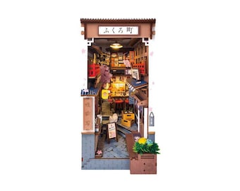 May include: A wooden model kit of a Japanese storefront with a sign that reads "ふくろう", which translates to "owl". The storefront has a small interior with a counter, shelves, and a cat sitting on a stool. The model is painted in a variety of colors, including brown, red, and white.