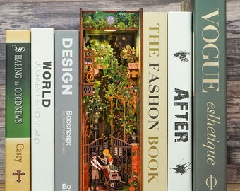 May include: A collection of books with various titles, including "Vogue" and "The Fashion Book." A detailed wooden book nook is placed between the books, featuring a miniature garden scene with figures. The books are arranged on a wooden surface.