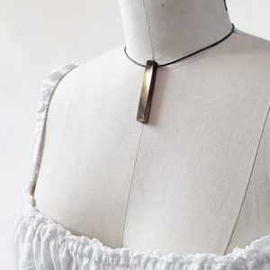 Può includere: Una collana semplice e minimalista con un ciondolo rettangolare lungo in legno marrone scuro. Il ciondolo è appeso a un sottile cordoncino nero.