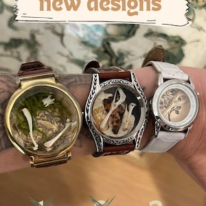 May include: Three unique wristwatches with transparent faces showcasing miniature art. The first watch has a gold case and brown leather strap, displaying bones and moss. The second watch has a silver case and brown strap, with a bone and animal figure. The third watch has a white strap and a floral design.
