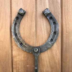May include: A rustic horseshoe-shaped hook with a metal finish. The hook is mounted on a wooden surface.