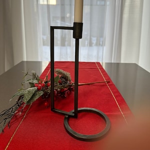 Peut inclure: Un bougeoir en métal noir avec une base carrée et un anneau circulaire au bas. Une bougie blanche est placée dans le bougeoir.
