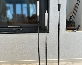 May include: Three black metal candle holders with white candles. The candle holders are tall and slender with a round base. The candles are lit and the flames are visible.