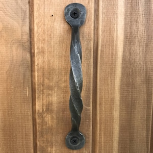 Puede incluir: Una manija de puerta de metal retorcido con un acabado gris oscuro. La manija est&aacute; unida a una puerta de madera con dos tornillos.