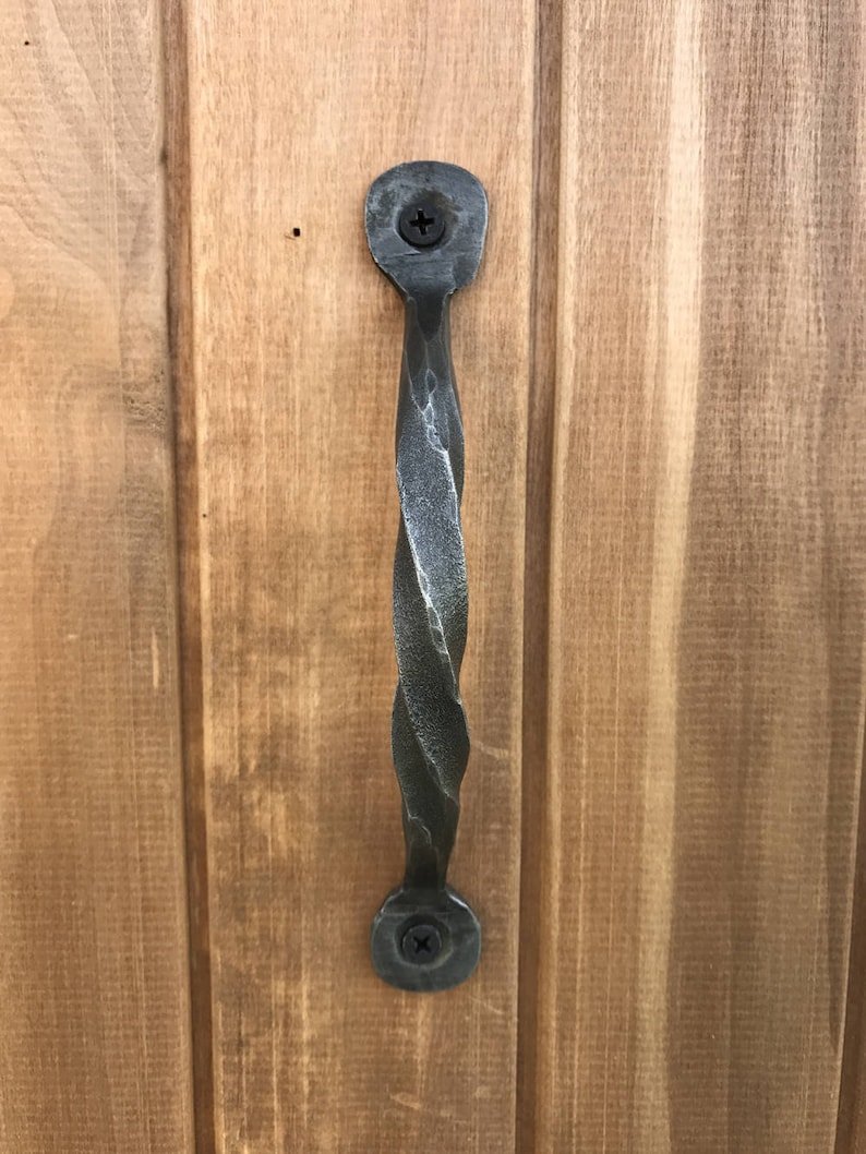 Puede incluir: Una manija de puerta de metal retorcido con un acabado gris oscuro. La manija est&aacute; unida a una puerta de madera con dos tornillos.