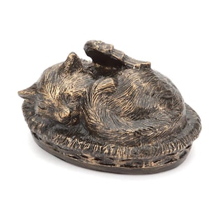 May include: A bronze-colored cat urn with a sleeping cat design. The cat is curled up in a bed with its head resting on its paws. The urn is oval shaped and has a detailed design.