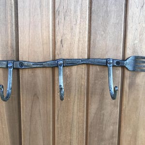 May include: A metal wall-mounted hook with three hooks and a fork design. The hook is made of a dark metal and has a rustic finish.