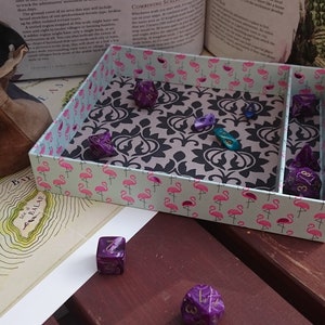 May include: A pink and black dice tray with a flamingo pattern. The tray has a divider to separate the dice. There are several purple and blue dice inside the tray.