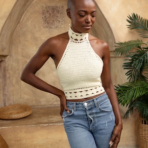 May include: A woman wearing a white crocheted halter top with a high neckline and a blue denim pair of jeans.