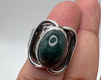 May include: A silver ring featuring a large, oval, dark green gemstone. The gemstone is set in a decorative silver setting with a floral or petal-like design. The ring is shown on a finger against a white background.