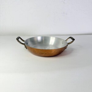 May include: A round, shallow copper pan with a polished silver-toned interior lining. The pan features two small, dark metal handles attached to opposite sides with visible rivets, providing a contrast to the warm, metallic exterior finish.
