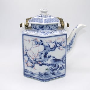 Könnte beinhalten: Eine weiße Porzellan-Teekanne mit einem blauen und rosa Blumendesign. Die Teekanne hat einen sechseckigen Körper und einen Deckel mit einem Messinggriff. Die Teekanne steht auf einem weißen Hintergrund.