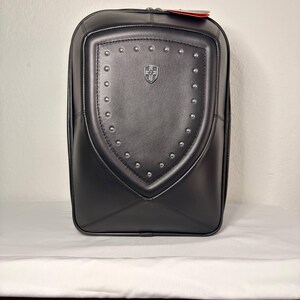 May include: Black backpack with a shield-shaped design on the front. The shield is adorned with silver studs and a small emblem. The backpack has a zipper closure and a red tag attached.