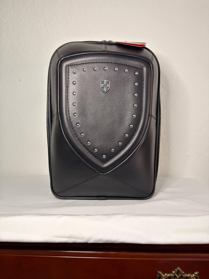 May include: Black backpack with a shield-shaped design on the front. The shield is adorned with silver studs and a small emblem. The backpack has a zipper closure and a red tag attached.