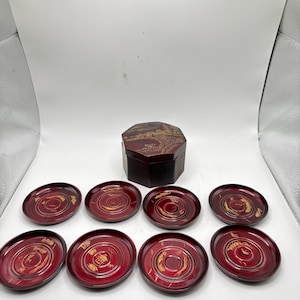 May include: A set of eight red and gold lacquered coasters with a matching hexagonal box. The coasters have a circular design with gold accents. The box features a landscape design on the lid.