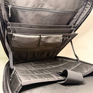 May include: Open view of a black briefcase with multiple compartments. The interior features several pockets, including zippered and open sections, and elastic straps. The lining has a subtle pattern. The briefcase is designed for organization and carrying documents and accessories.