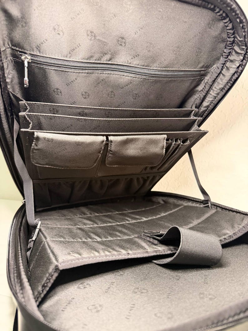 May include: Open view of a black briefcase with multiple compartments. The interior features several pockets, including zippered and open sections, and elastic straps. The lining has a subtle pattern. The briefcase is designed for organization and carrying documents and accessories.
