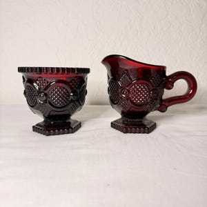 Könnte beinhalten: Ein rubinrotes Sahne- und Zuckerdosen-Set aus Glas. Der Sahnebereiter hat einen Griff und einen Ausguss, während die Zuckerdose offen ist. Beide Teile weisen ein Diamanten- und Kreismuster auf und stehen auf einem Sockel. Das Set ist wahrscheinlich Vintage.