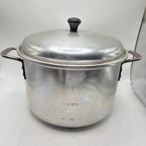 Op de afbeelding: Een zilveren metalen kookpot met een deksel en twee handvatten. De pot heeft een zwarte knop op het deksel en meetmarkeringen voor 1, 2 en 3 liter. De pot is ontworpen om te koken en voedsel te bereiden.