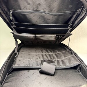 May include: Black leather backpack with multiple interior compartments and pockets. The interior lining features a repeating pattern. The backpack has a zippered main compartment and several organizational sections for documents and accessories.