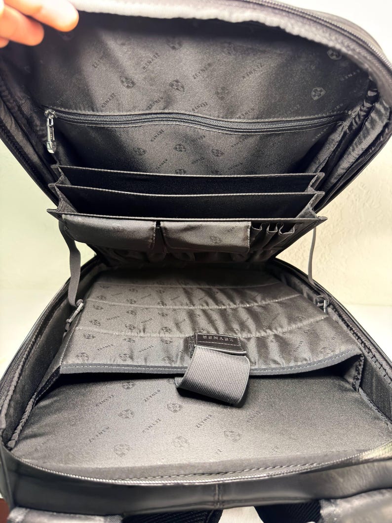 May include: Black leather backpack with multiple interior compartments and pockets. The interior lining features a repeating pattern. The backpack has a zippered main compartment and several organizational sections for documents and accessories.