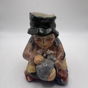 May include: A ceramic figurine of a seated figure wearing a green hat and brown coat, holding a gray sack. The figurine has a detailed face and is painted with various colors.
