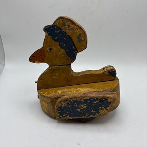 May include: A vintage wooden duck toy with a yellow body, a black and blue hat, and an orange beak. The toy has a distressed, worn appearance, suggesting it is an antique or a well-loved item. The duck is a classic children's toy.
