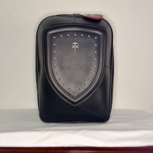 May include: Black leather backpack with a shield-shaped design on the front, adorned with silver studs and a small emblem. The backpack has a rectangular shape with rounded corners and a zipper closure. A red tag hangs from the top.