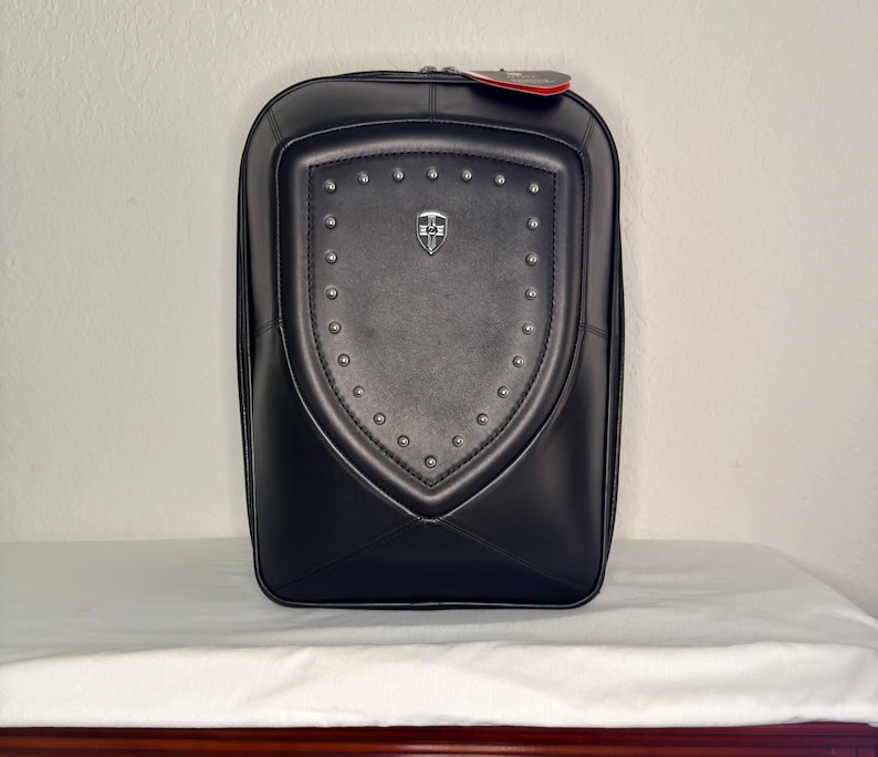 May include: Black leather backpack with a shield-shaped design on the front, adorned with silver studs and a small emblem. The backpack has a rectangular shape with rounded corners and a zipper closure. A red tag hangs from the top.