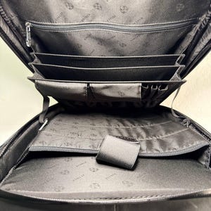 May include: Open view of a black backpack with multiple compartments. The interior features a zippered pocket, several card slots, and a large open space. The lining has a repeating pattern of a logo. The backpack is designed for organization and carrying essentials.