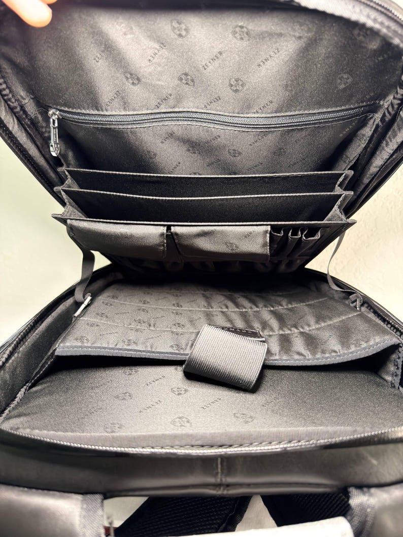May include: Open view of a black backpack with multiple compartments. The interior features a zippered pocket, several card slots, and a large open space. The lining has a repeating pattern of a logo. The backpack is designed for organization and carrying essentials.