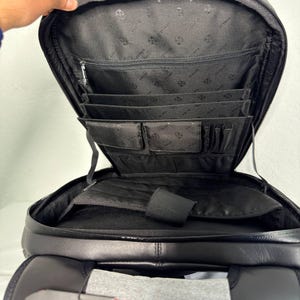 May include: Black leather backpack with multiple interior compartments, including pockets and dividers. The backpack is open, revealing the interior lining and organizational features. The top is open, and the zipper is visible. The bag is designed for carrying various items.