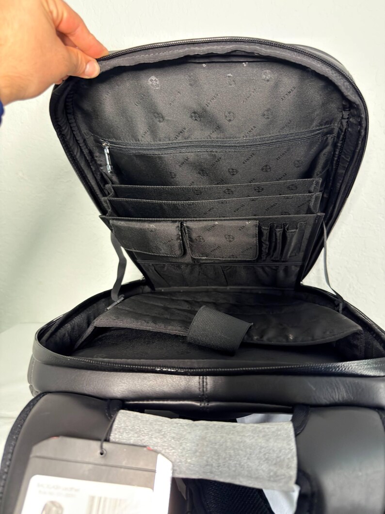 May include: Black leather backpack with multiple interior compartments, including pockets and dividers. The backpack is open, revealing the interior lining and organizational features. The top is open, and the zipper is visible. The bag is designed for carrying various items.