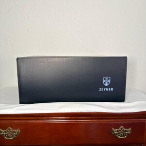 May include: A black rectangular box with the word "ZEYNER" printed in white. The box has a shield logo on the right side. The box is sitting on a white cloth on top of a wooden dresser.