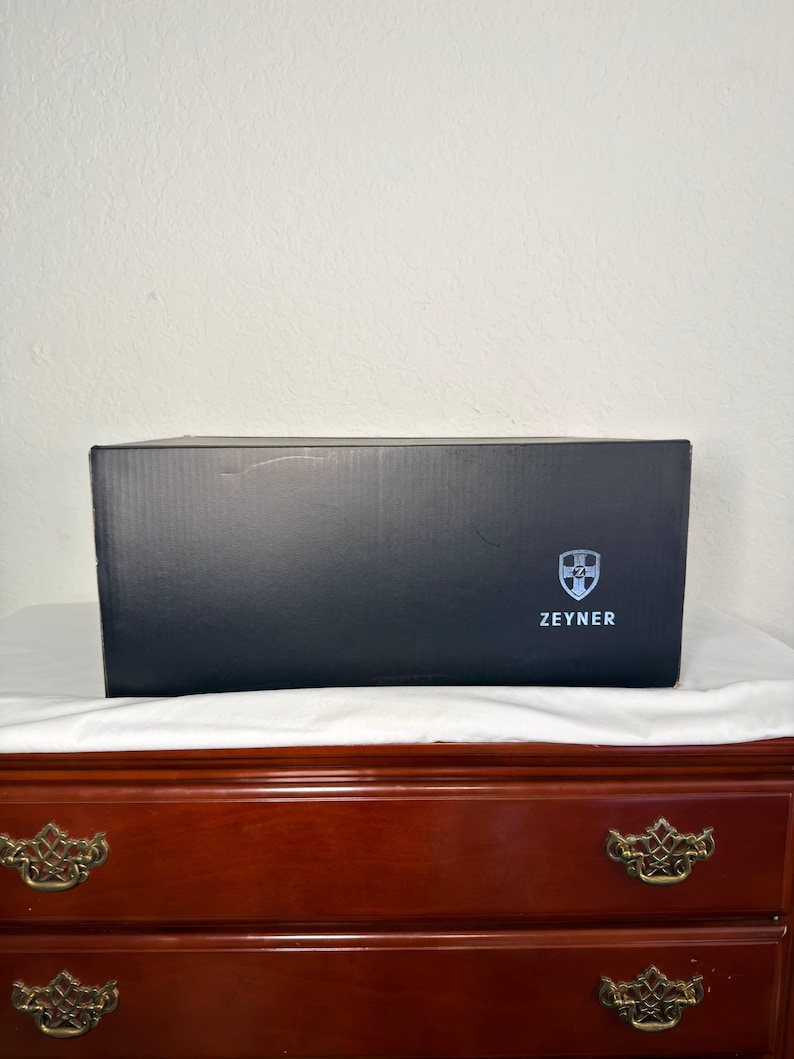 May include: A black rectangular box with the word "ZEYNER" printed in white. The box has a shield logo on the right side. The box is sitting on a white cloth on top of a wooden dresser.
