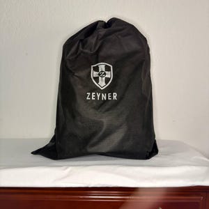 May include: A black drawstring bag with the white text "ZEYNER" and a shield logo. The bag is sitting on a white surface, and the background is a plain white wall. The bag is made of a soft, matte material.