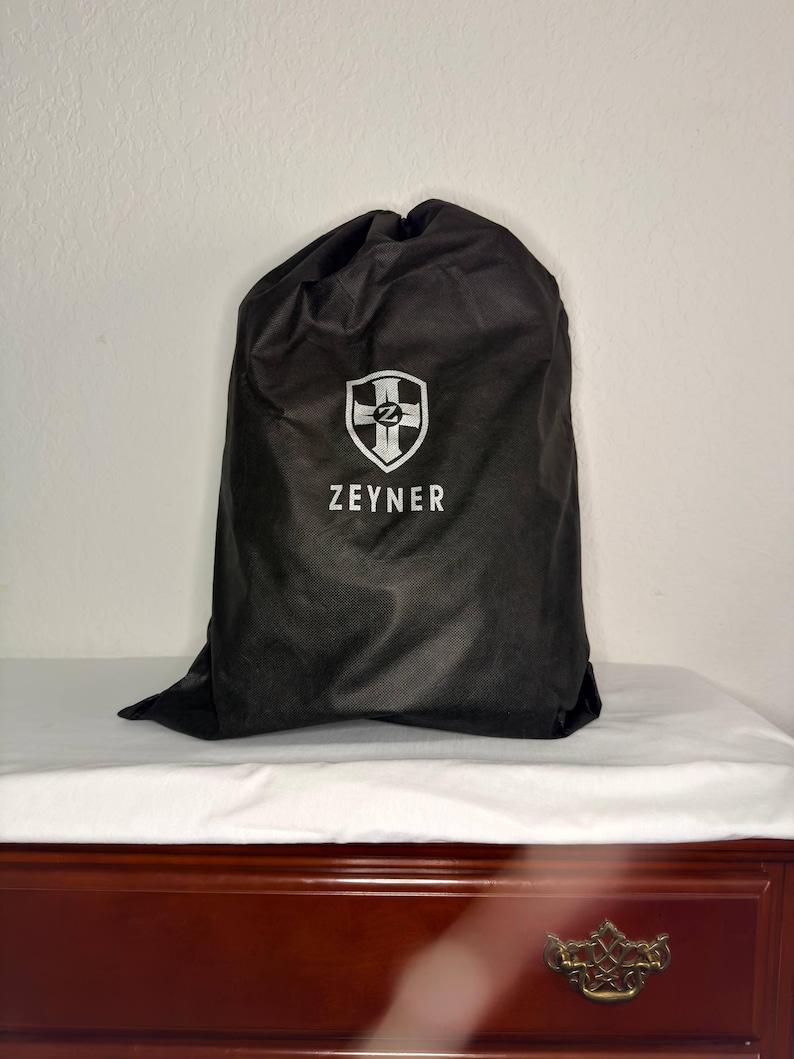 May include: A black drawstring bag with the white text "ZEYNER" and a shield logo. The bag is sitting on a white surface, and the background is a plain white wall. The bag is made of a soft, matte material.