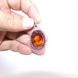 Puede incluir: Un colgante de plata con una piedra de ámbar facetada con un diseño de camafeo. El colgante tiene un borde de cuerda.